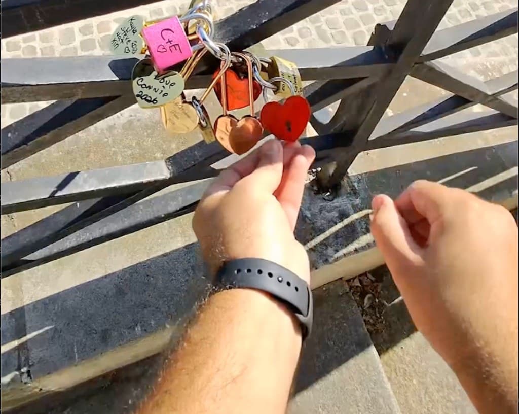 A red heart padlock being locked by hand on the Pont des Arts railing, among other love locks